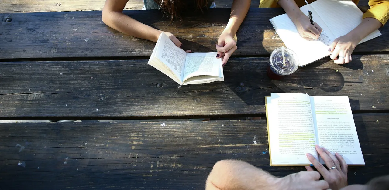 Hands hold books and write in a notebook around a sunlit wooden table.