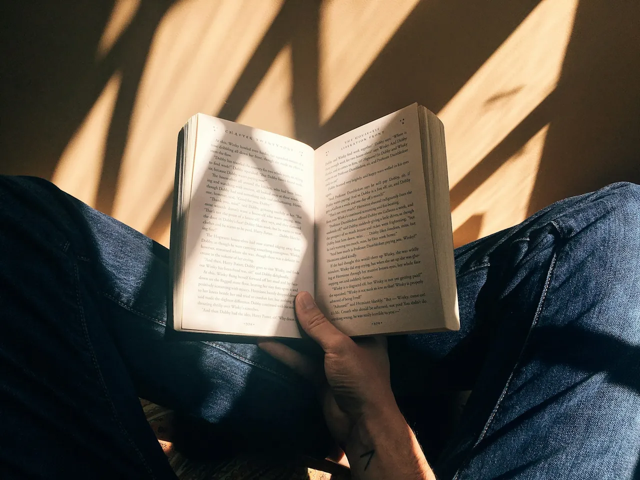 A hand holds an open book over blue jeans in warm sunlight and window shadows.